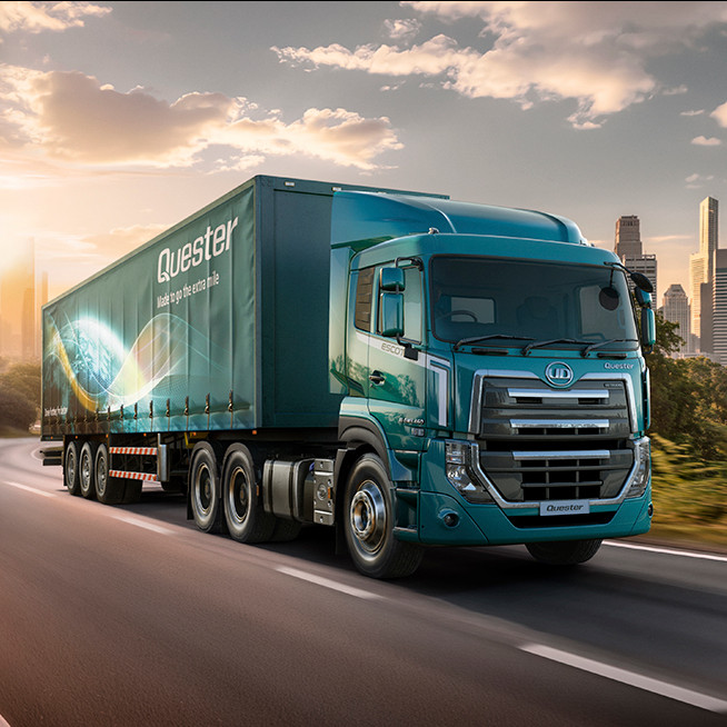 Blue semi-truck driving on a scenic road with city skyline in the background at sunset.