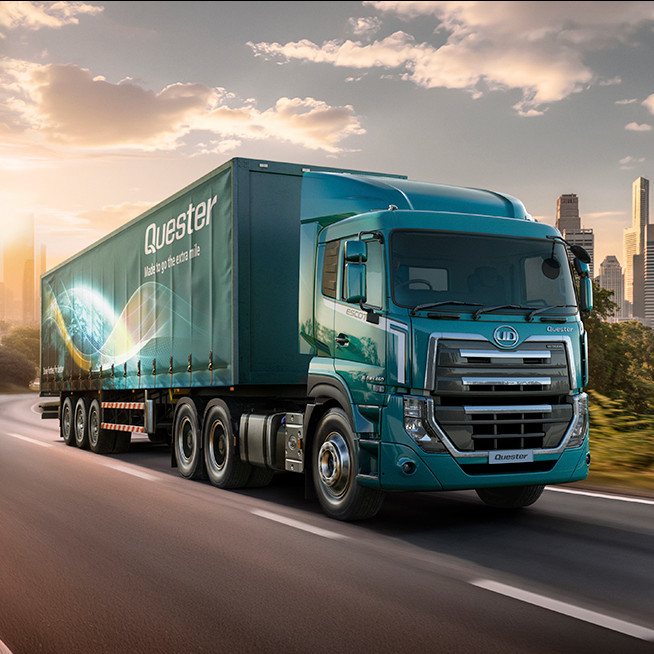 Blue semi-truck driving on a highway with a city skyline in the background at sunset.