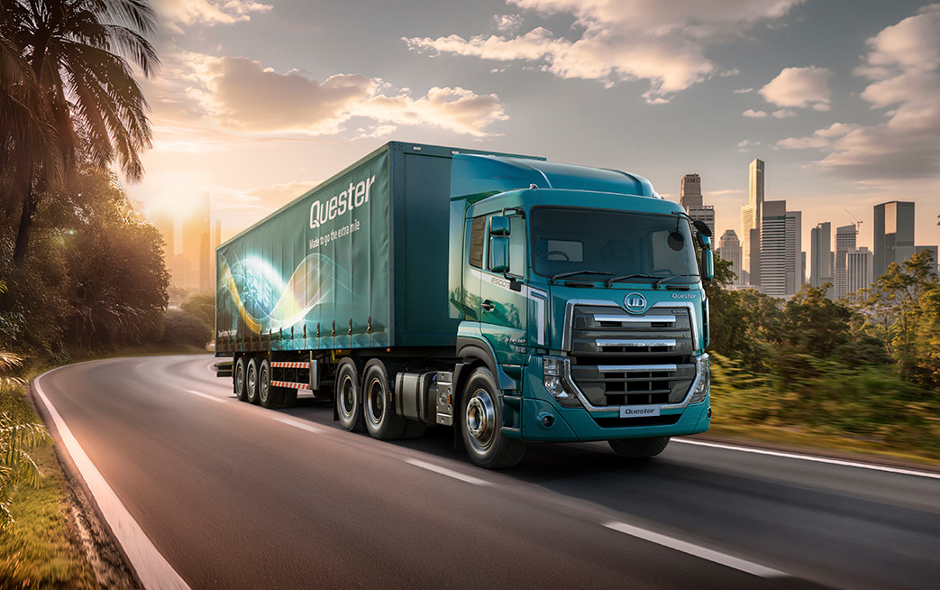 Teal semi-truck driving on a road with a city skyline in the background at sunset.