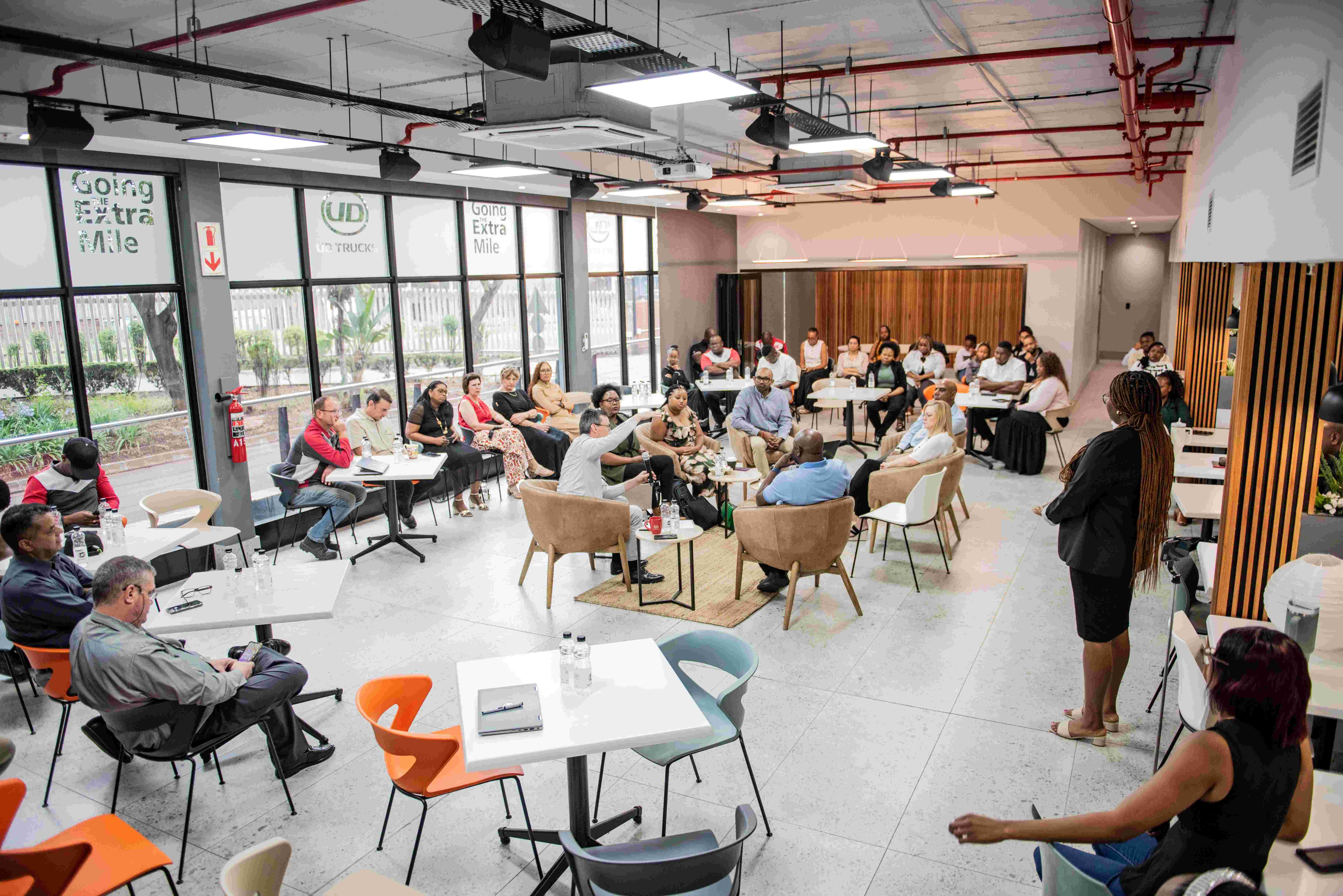 Mense sit in ‘n moderne vergaderlokaal en luister na ‘n spreker voor in die kamer.