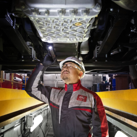 Mechanic with helmet inspects car underside using flashlight in workshop.