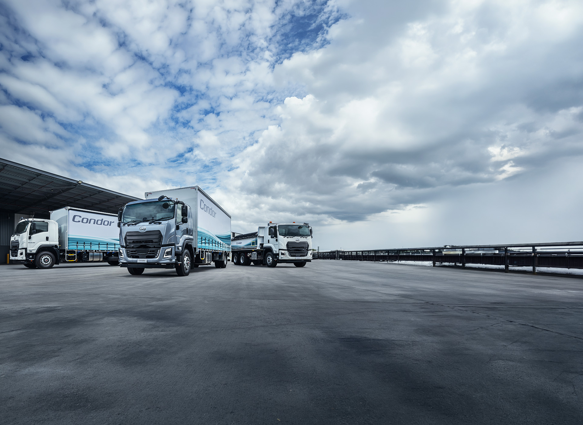 Three delivery trucks parked on a wet asphalt lot under a cloudy sky.