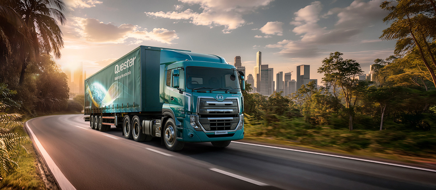 Teal semi-truck driving on a road with a city skyline in the background at sunset.