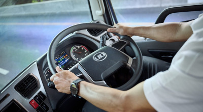 Person driving a commercial truck, hands on the steering wheel, dashboard visible.