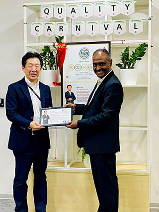 दो पुरुष सूट में प्रमाणपत्र देते हुए, पीछे पौधे और "QUALITY CARNIVAL" बोर्ड.